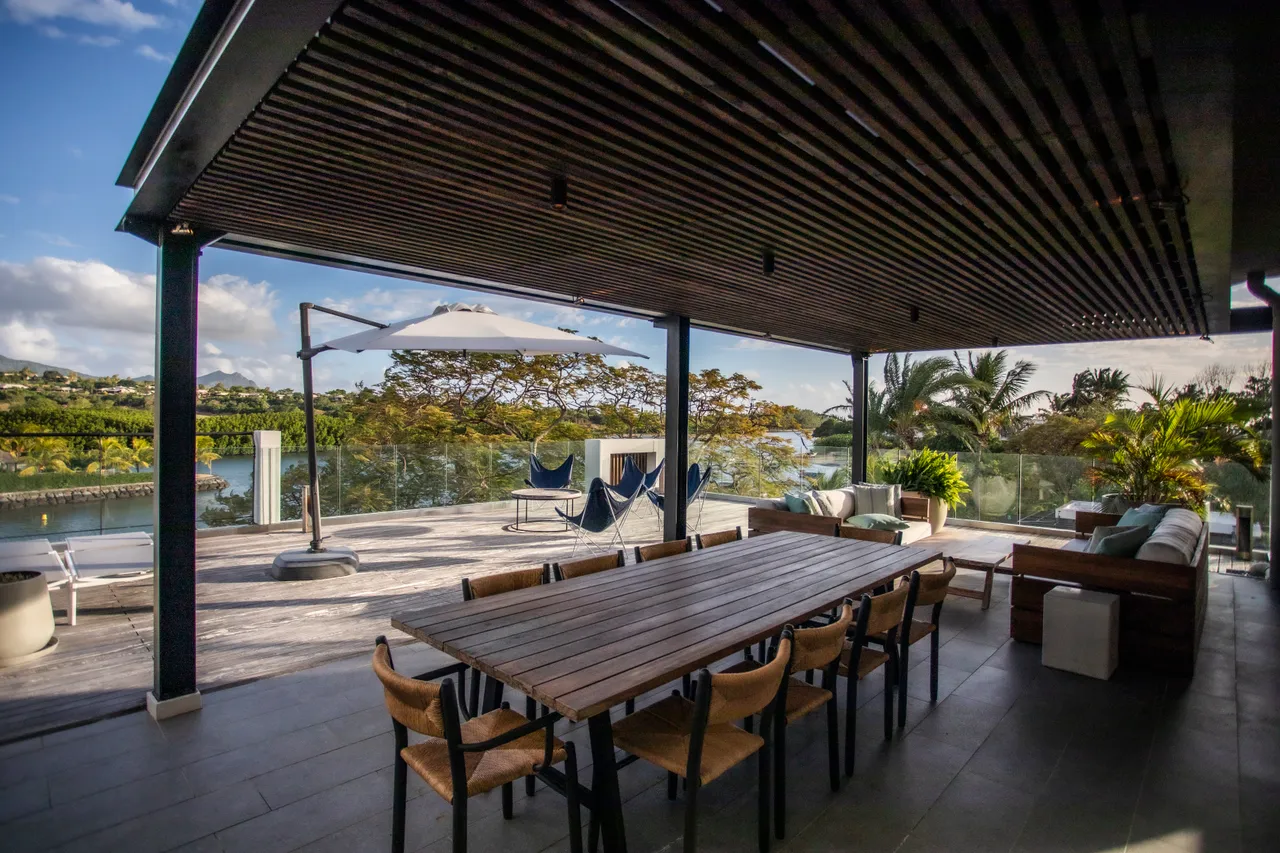Bois foncé du plafond créant une atmosphère chaleureuse en extérieur. Grande table en bois offrant un espace convivial pour les repas. Chaises confortables disposées autour de la table. Vue dégagée sur la nature environnante ajoutant une dimension apaisante. Canapés en extérieur favorisant la détente.