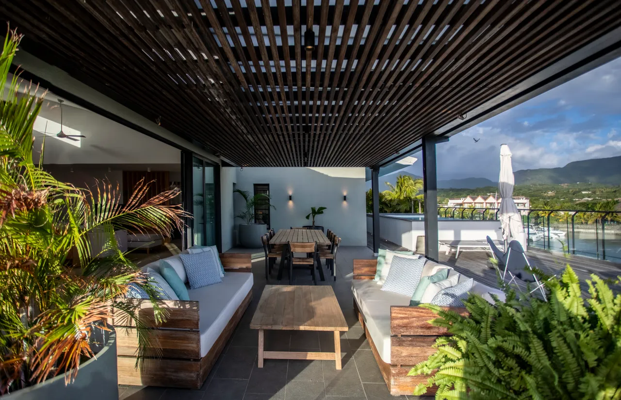 Plafond en bois avec éclairage intégré créant une ambiance chaleureuse. 

Espace de vie ouvert offrant de confortables canapés et tables en bois. 

Végétation soigneusement choisie ajoutant une touche naturelle à l'aménagement. 

Zone de repas extérieure équipée de grandes assises pour partager des repas. 

Grands espaces vitrés permettant une vue sur l'extérieur et
