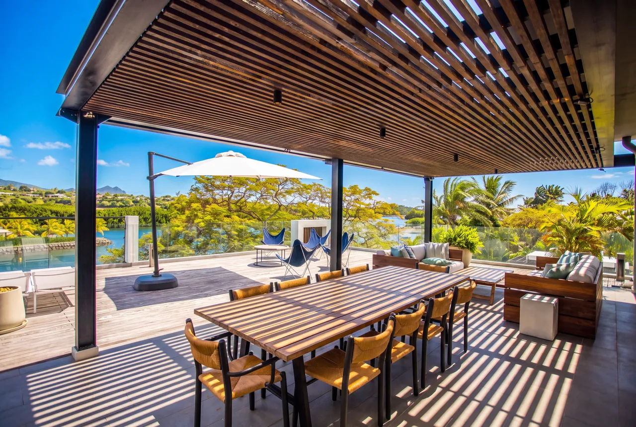 Espace repas sous une pergola à lames, avec table longue et vue sur la marina.