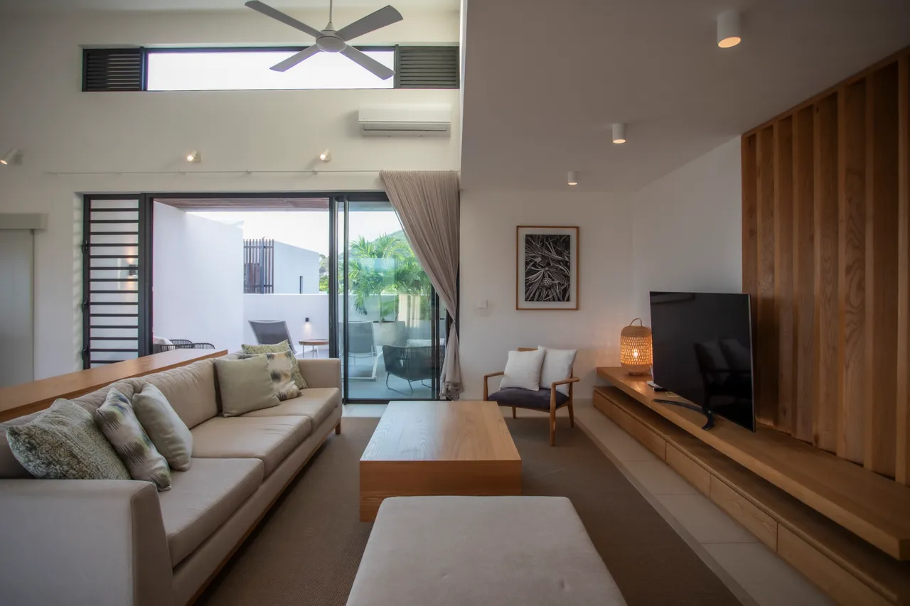 Lumière naturelle se diffusant à travers de grandes baies vitrées. 

Espace de vie moderne avec un mobilier en bois chaleureux. 

Coin salon confortable avec un canapé spacieux et des coussins. 

Écran de télévision placé sur un meuble en bois sobre. 

Accès direct à la terrasse depuis le salon, favorisant la circulation.