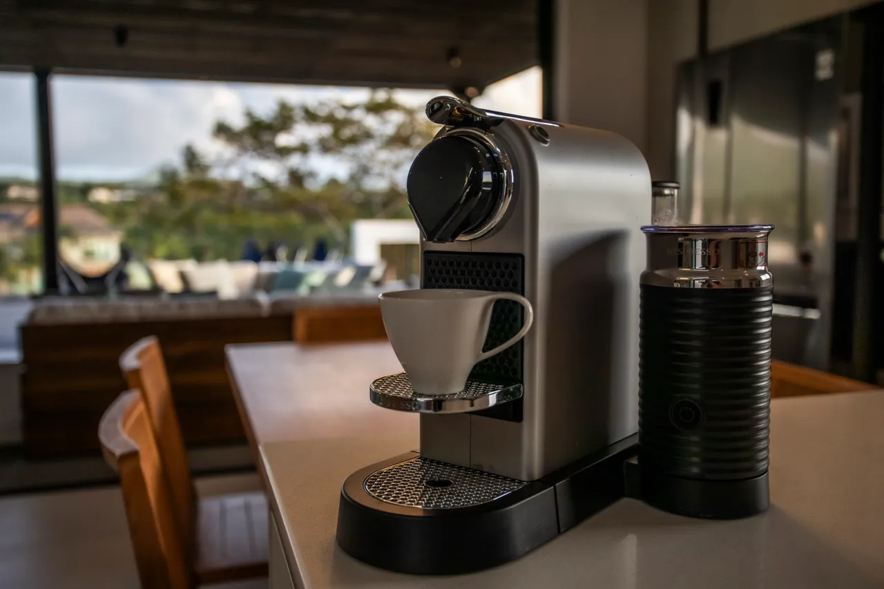 Machine à café moderne sur un comptoir en marbre. 

Mobilier en bois clair créant une ambiance chaleureuse et accueillante. 

Espace lumineux avec grandes fenêtres offrant une vue sur l'extérieur. 

Coin repas à proximité, idéal pour des repas conviviaux. 

Plan de travail spacieux pour faciliter les préparations culinaires.