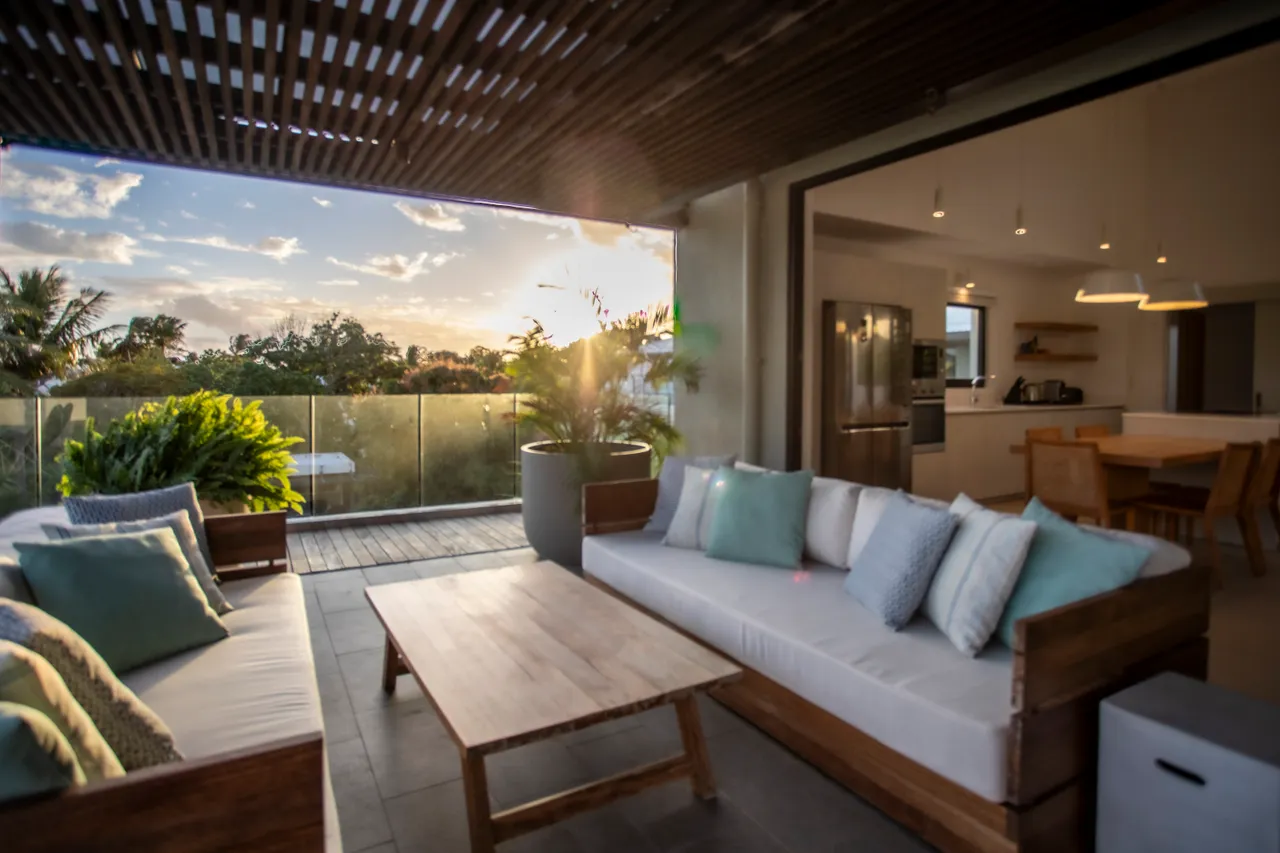 Espace extérieur avec salon confortable et vue sur la végétation. 

Mobilier en bois clair, favorisant une ambiance naturelle et accueillante. 

Lumière naturelle filtrant à travers le plafond en lattes pour une atmosphère agréable. 

Accès direct à la cuisine et coin repas depuis la terrasse. 

Éclairage suspendu ajoutant une touche moderne à l'espace.
