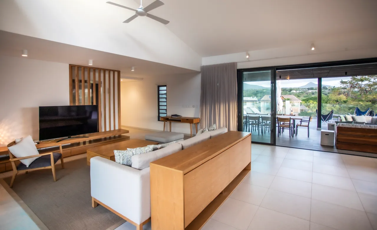 Sol carrelé clair assurant une ambiance moderne et épurée. 

Mobilier en bois combiné avec des pièces contemporaines pour un agencement harmonieux. 

Large baie vitrée permettant un accès direct à la terrasse extérieure. 

Espace de vie spacieux avec coin salon bien défini. 

Éclairage intégré offrant une atmosphère agréable et conviviale.