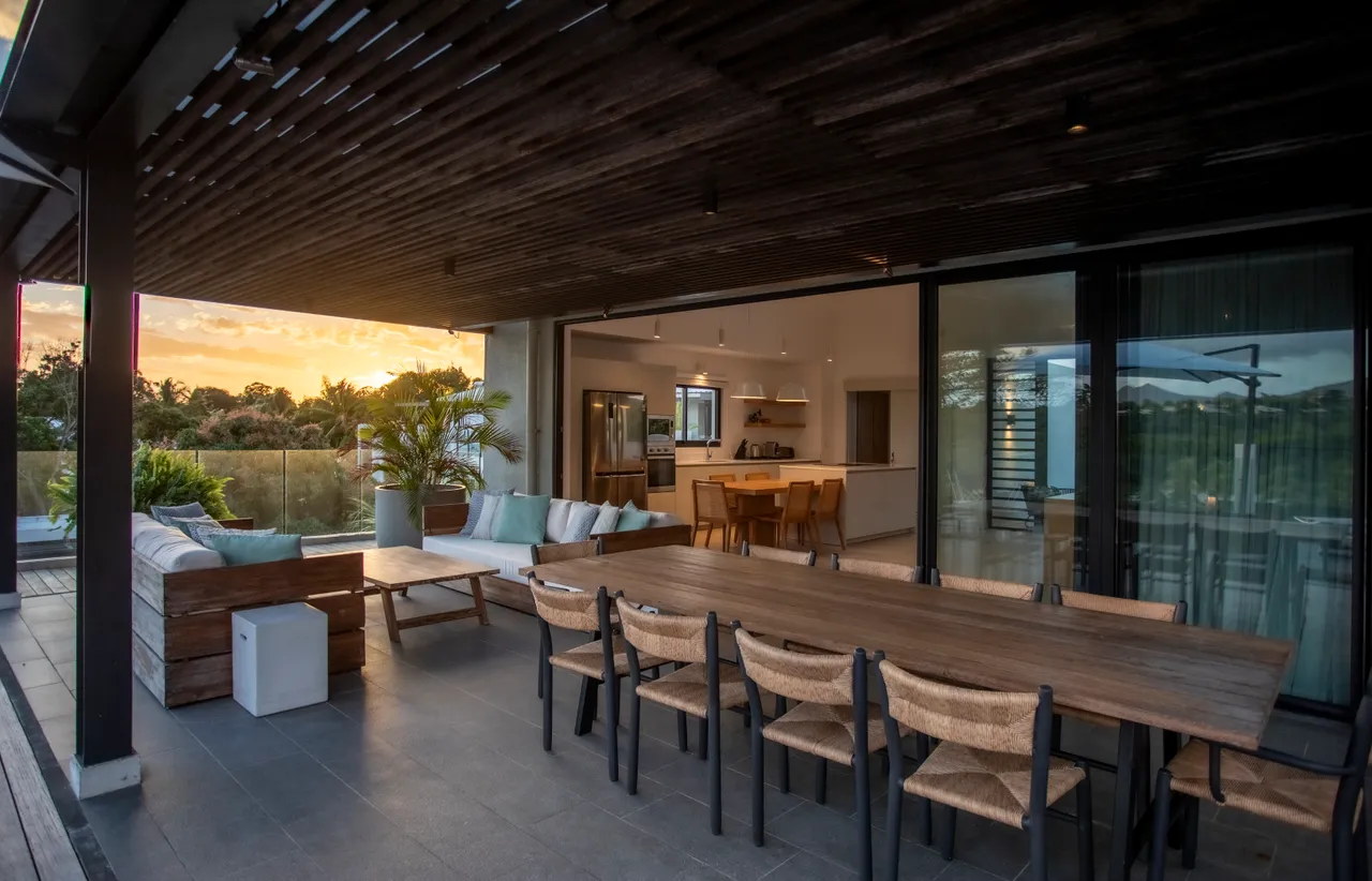 Espace extérieur avec coin repas en bois et vue dégagée.  
Terrasse couverte avec canapés et végétation luxuriante.  
Cuisine moderne ouverte sur la salle à manger.  
Chaises en osier autour de la table à manger.  
Ambiance chaleureuse grâce à l'éclairage suspendu.  
Grandes baies vitrées pour une luminosité optimale.  
É