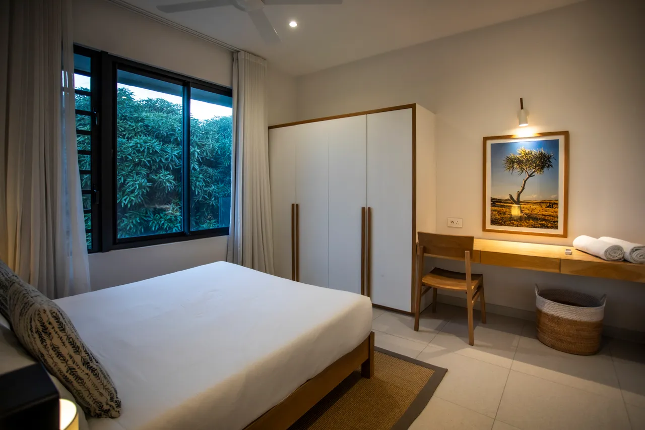 Chambre avec lit king-size et grandes fenêtres donnant sur la végétation. 

Espace de rangement intégré permettant un agencement fonctionnel. 

Coin bureau en bois offrant un espace de travail pratique. 

Linge de lit coordonné apportant une touche de confort. 

Éclairage chaleureux créant une atmosphère reposante.