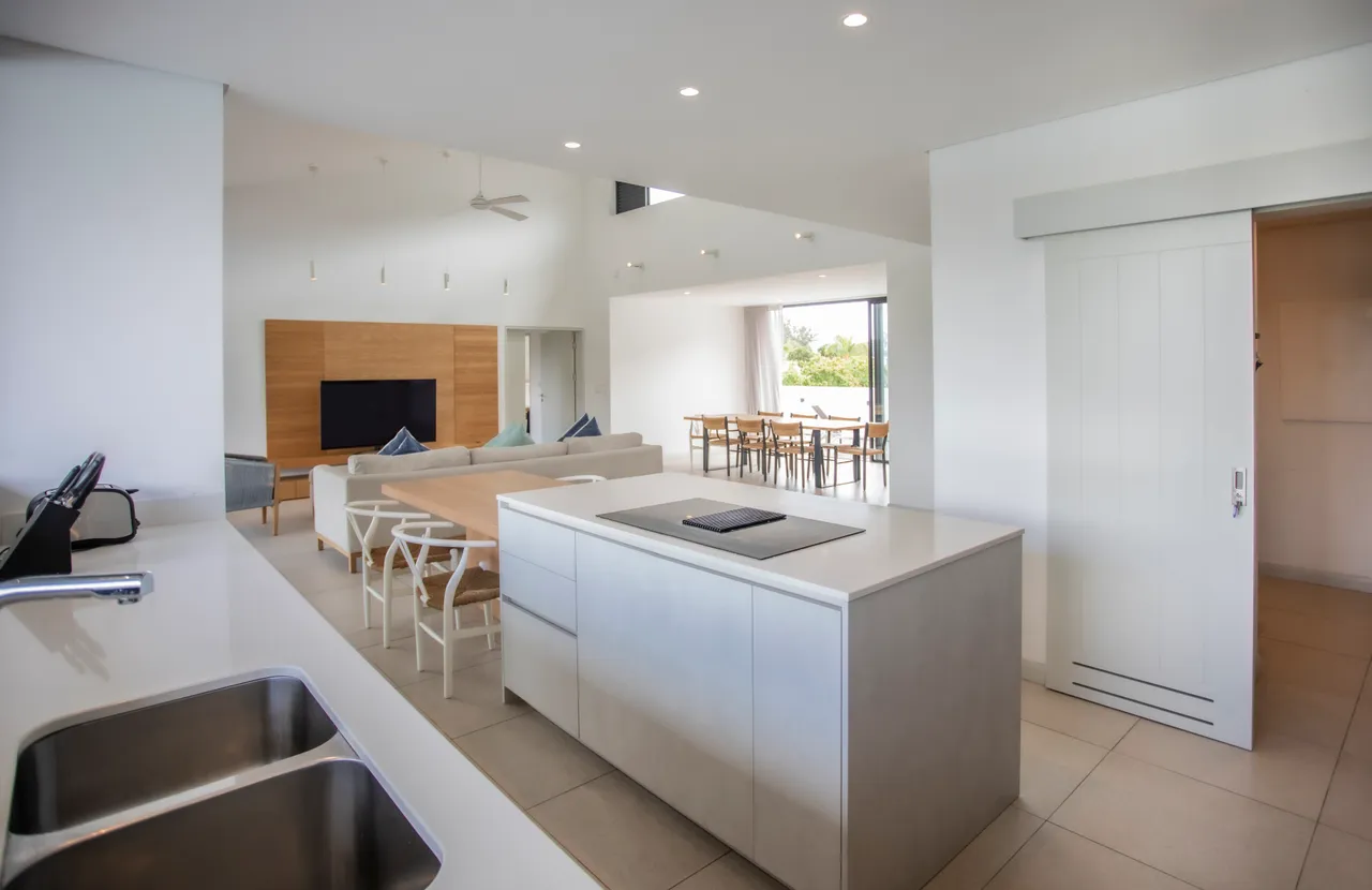 Cuisine ouverte avec plan de travail en béton et espace de rangement pratique.  
Mobilier clair et fonctionnel intégrant un espace pour les repas.  
Lumière naturelle abondante grâce aux grandes fenêtres sur l'extérieur.  
Sol en carrelage clair apportant une ambiance aérienne à l'espace.  
Accès direct à l'espace de vie depuis la cuisine pour un agencement fluide.