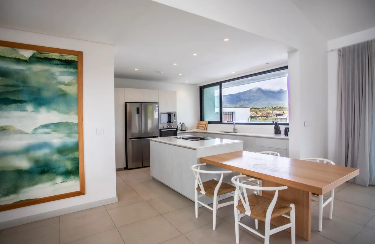 Cuisine moderne avec un plan de travail clair et un espace de rangement fonctionnel.  
Lumière naturelle pénétrant à travers la grande fenêtre offrant une vue sur la montagne.  
Équipements intégrés facilitant la préparation des repas au quotidien.  
Mobilier simple avec des chaises en bois autour de la table à manger.  
Sol en carrelage clair assurant une ambiance aérée