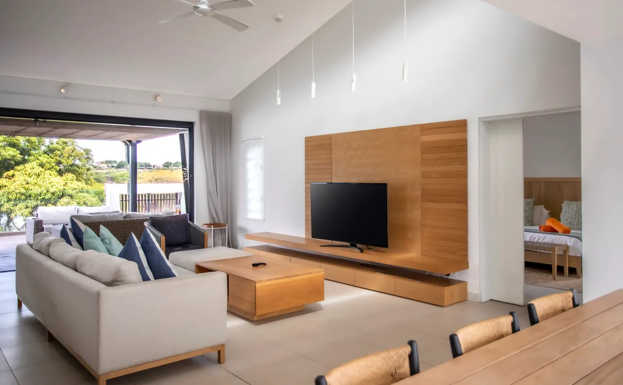 Sol en carrelage clair créant une ambiance aérée dans l'espace de vie.  
Mobilier en bois intégré pour un agencement harmonieux et fonctionnel.  
Lumière naturelle abondante grâce aux larges baies vitrées sur l'extérieur.  
Téléviseur monté sur un mur en bois pour une esthétique contemporaine.  
Accès direct à la terrasse depuis le salon
