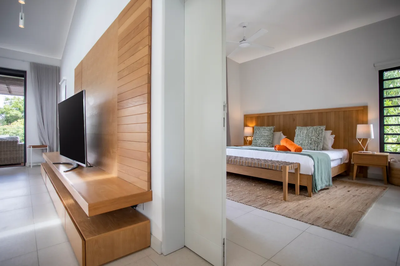 Chambre équipée d'un lit en bois et d'un espace de rangement pratique.  
Grandes fenêtres offrant une vue sur la nature environnante depuis l'intérieur.  
Coin salon adjacent avec accès direct à la terrasse extérieure.  
Mobilier minimaliste favorisant une ambiance apaisante et fonctionnelle.  
Tapis en fibres naturelles ajoutant une texture douce au sol.