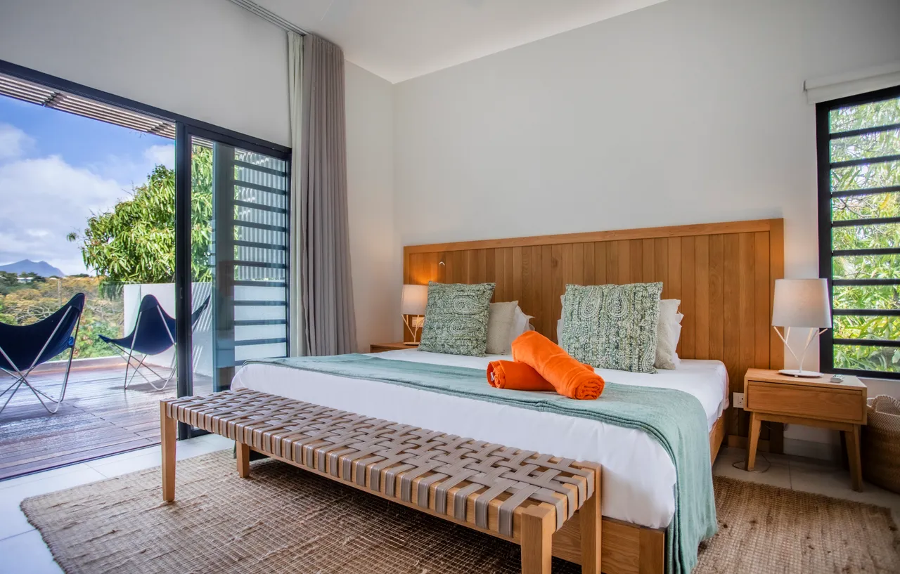 Chambre avec accès direct à la terrasse extérieure et vue sur la nature.  
Lit spacieux avec têtes de lit en bois créant un cadre accueillant.  
Éléments décoratifs simples ajoutent une touche harmonieuse à l'espace.  
Lumière naturelle généreuse grâce aux grandes fenêtres favorisant un environnement lumineux.  
Tapis en fibres naturelles apportant une texture douce au sol