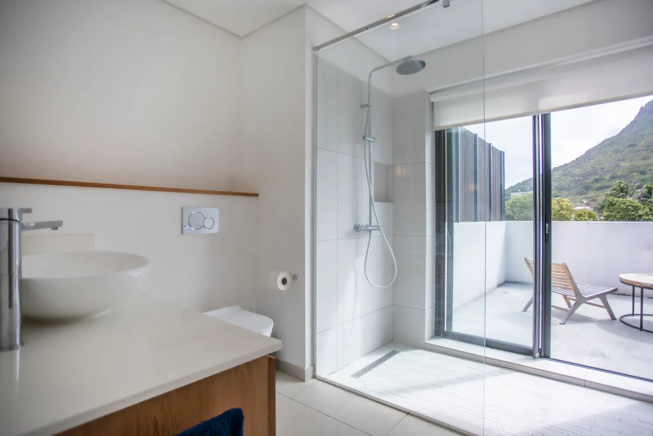 Salle de bains avec douche italienne et accès à une petite terrasse.  
Éclairage naturel grâce à une fenêtre offrant une vue sur l'extérieur.  
Plan de travail clair et lavabo surmonté d'un meuble en bois.  
Mobilier fonctionnel et sobre créant un espace pratique et aéré.  
Sol en carrelage beige apportant une cohérence à l'esthétique.