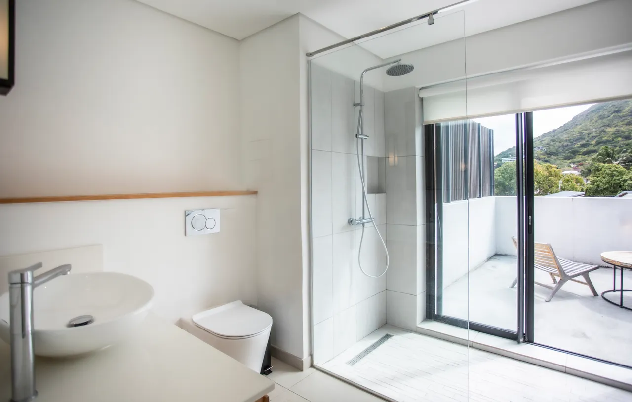 Salle de bains moderne avec douche italienne et éclairage naturel.  
Lavabo sur plan de travail clair, intégrant un meuble de rangement.  
Sol en carrelage beige assurant une ambiance lumineuse et aérée.  
Fenêtre offrant une vue sur la terrasse adjacente.  
Mobilier fonctionnel et sobre favorisant une atmosphère pratique.