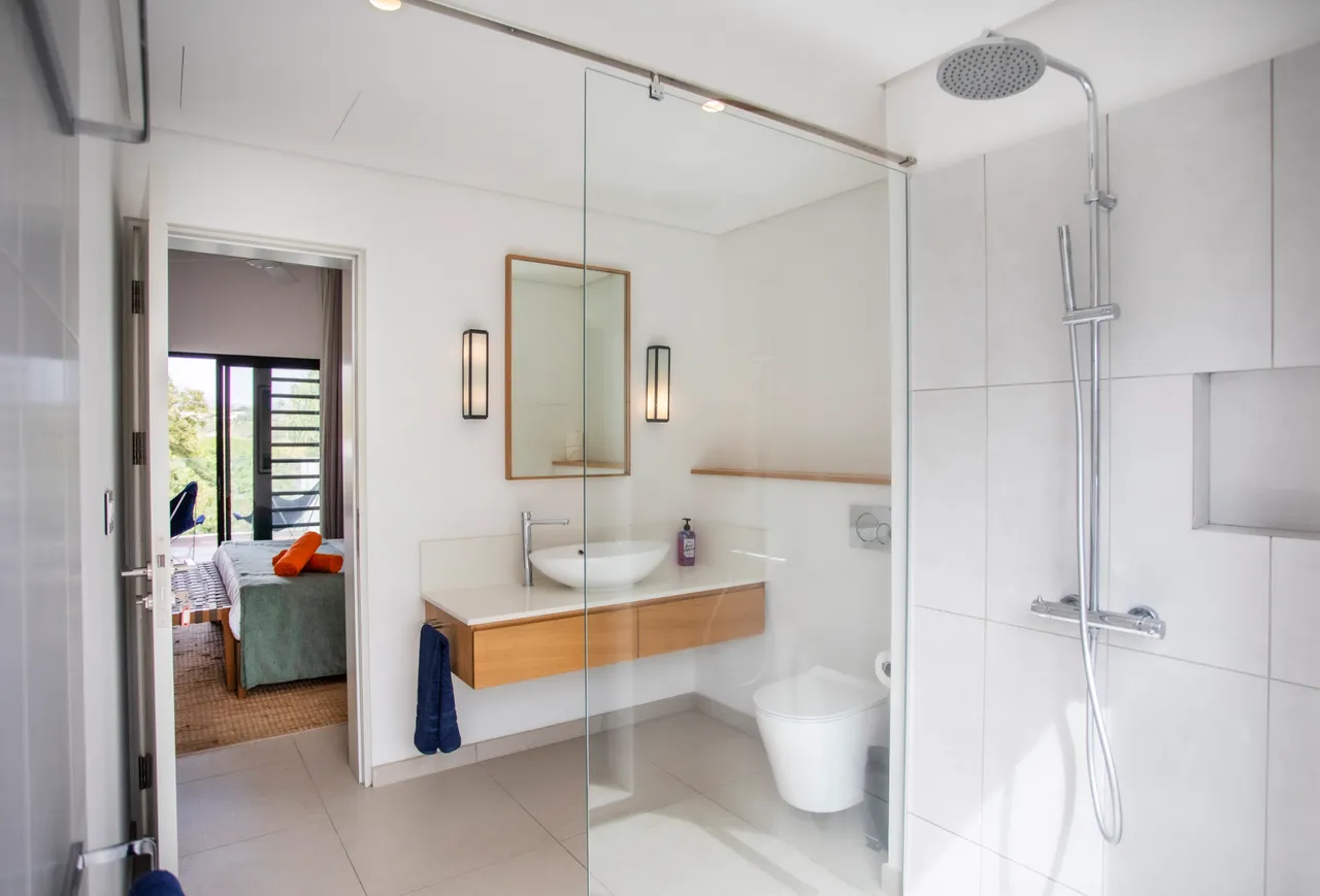 Salle de bains équipée d'une douche italienne et de rangements pratiques.  
Lumière naturelle entrant par une fenêtre sur l'extérieur.  
Plan de travail en bois clair offrant un espace fonctionnel.  
Détails minimalistes assurant une ambiance épurée et contemporaine.  
Sol en carrelage clair ajoutant une touche de légèreté à l'ensemble.