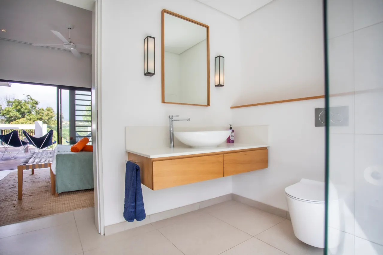 Salle de bains moderne avec douche italienne et espace de rangement intégré.  
Lumière naturelle apportée par une fenêtre offrant une vue dégagée.  
Plan de travail clair avec lavabo suspendu pour un design épuré.  
Éclairage minimaliste soulignant l'aspect fonctionnel de l'espace.  
Sol en carrelage clair créant une atmosphère lumineuse.