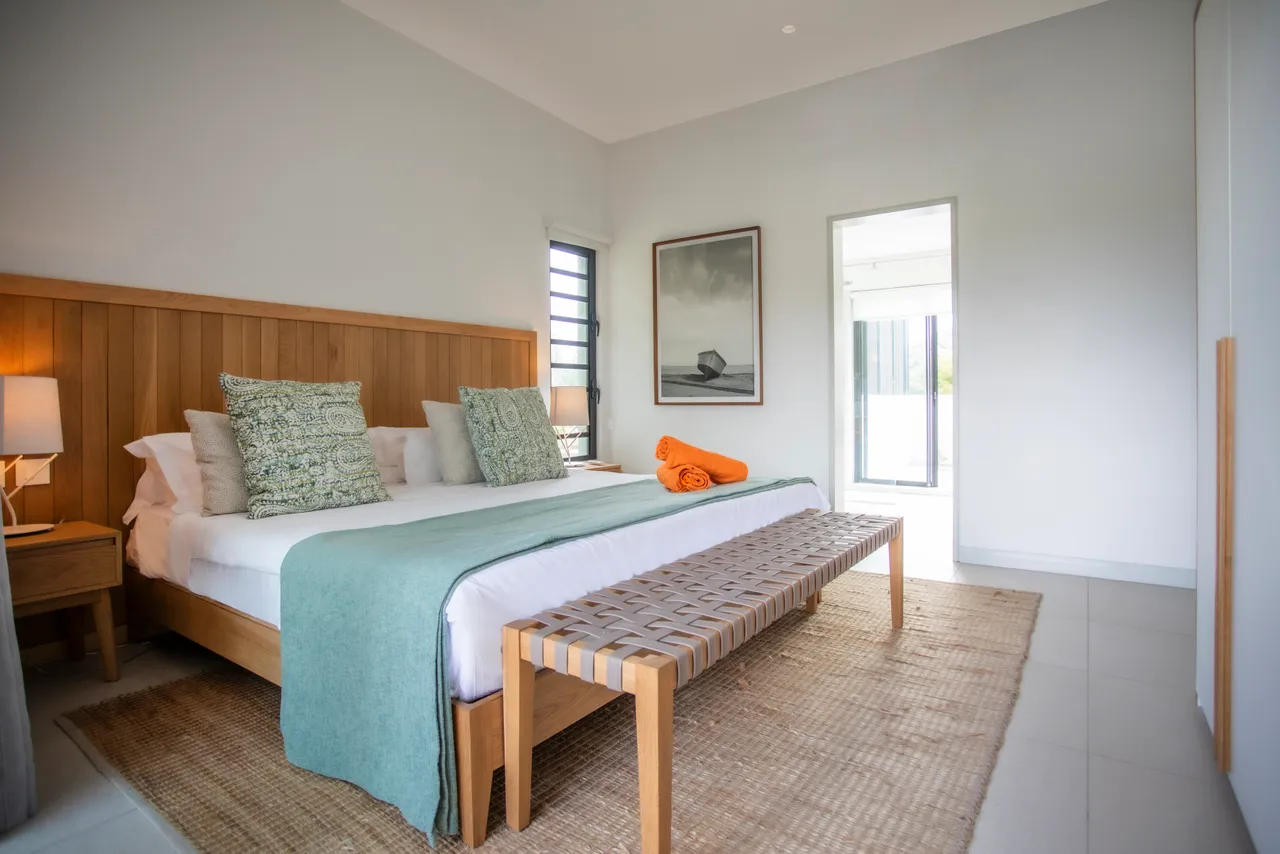 Chambre spacieuse avec accès direct à la terrasse et décoration épurée.  
Lit en bois avec têtes de lit intégrées et coussins colorés.  
Éléments naturels comme le tapis en jute ajoutant de la texture.  
Fenêtres offrant une vue dégagée sur l'extérieur et la végétation.  
Mobilier fonctionnel incluant un espace de rangement optimisé.