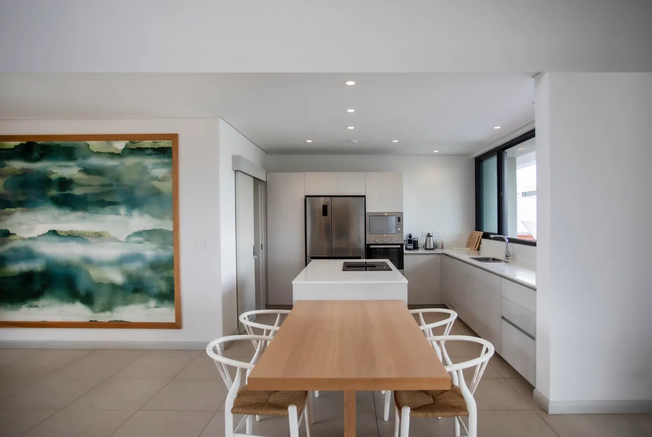 Tons neutres et textures douces dans la cuisine moderne.  
Espace fonctionnel avec îlot central et électroménagers intégrés.  
Larges fenêtres permettant une bonne luminosité naturelle.  
Mobilier minimaliste offrant un agencement pratique pour les repas.  
Table en bois claire ajoutant une note chaleureuse à l'ensemble.