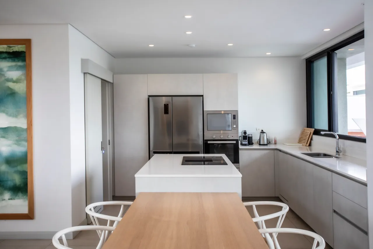 Cuisine contemporaine équipée d'un îlot central et de rangements intégrés.  
Électroménagers modernes offrant un agencement fonctionnel pour la préparation des repas.  
Lumière naturelle abondante grâce aux grandes fenêtres en façade.  
Mobilier minimaliste en bois pour une ambiance épurée et accueillante.  
Table en bois claire créant un espace pratique pour les repas.