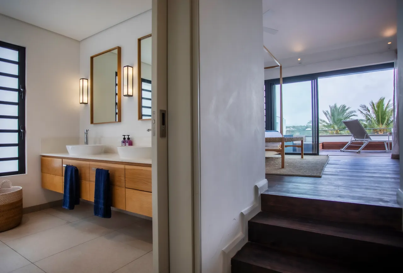 Salle de bains moderne avec double vasque et rangements intégrés.  
Miroirs encadrés apportant une dimension supplémentaire à l'espace.  
Lumière naturelle filtrée par de grandes fenêtres offrant une vue extérieure.  
Décoration épurée soulignant l'aspect fonctionnel du lieu.  
Accès direct à la chambre depuis l'espace sanitaire.
