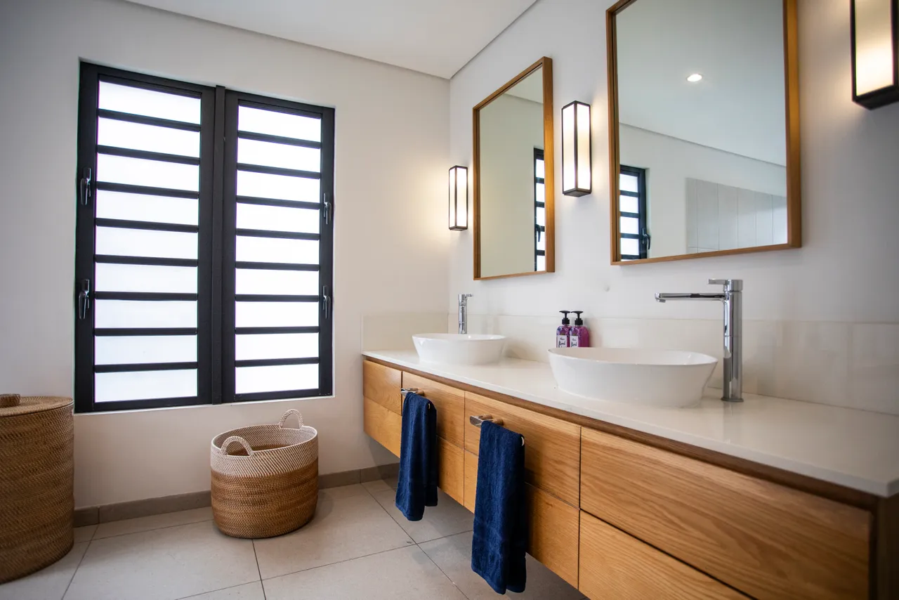 Salle de bains lumineuse avec deux lavabos et rangements intégrés.  
Miroirs encadrés apportant une profondeur à l'espace.  
Fenêtres offrant une lumière naturelle agréable tout en préservant l'intimité.  
Décoration sobre mettant en avant la fonctionnalité des éléments.  
Accès direct à la chambre via un couloir pratique.