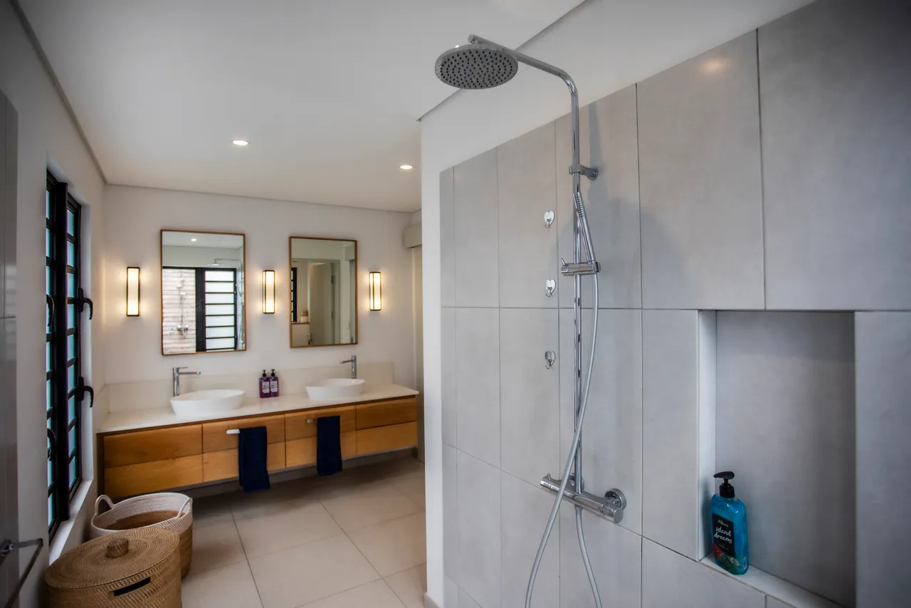 Teintes claires et surfaces lisses dans la salle de bains.  
Double vasque encadrée par des miroirs augmentant la luminosité.  
Douche spacieuse équipée d'une paroi en verre pour un accès facile.  
Rangements intégrés optimisant l’espace disponible pour les produits.  
Fenêtres stratégiquement placées assurant lumière naturelle et intimité.