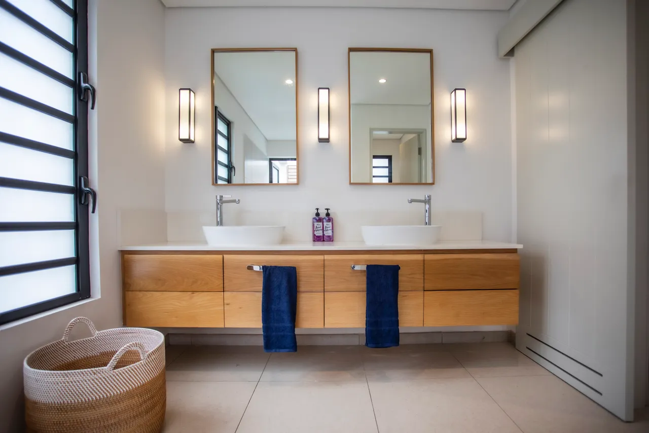 Salle de bains avec mobilier en bois et doubles vasques.  
Miroirs lumineux créant une sensation d'espace et de clarté.  
Sols en carrelage clair apportant une touche contemporaine.  
Éclairage intégré soulignant l'aspect moderne de la pièce.  
Rangements pratiques optimisant l'organisation des produits de beauté.