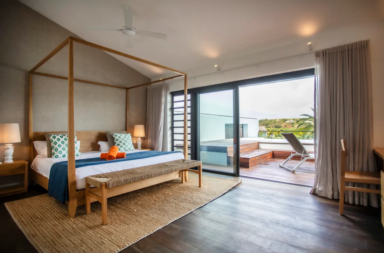 Chambre avec accès direct à une terrasse en bois.  
Lit à baldaquin ajoutant une touche de confort.  
Grandes fenêtres offrant une vue sur l'extérieur.  
Mobilier en bois clair pour une atmosphère apaisante.  
Tapis en jute apportant une texture naturelle à l'espace.
