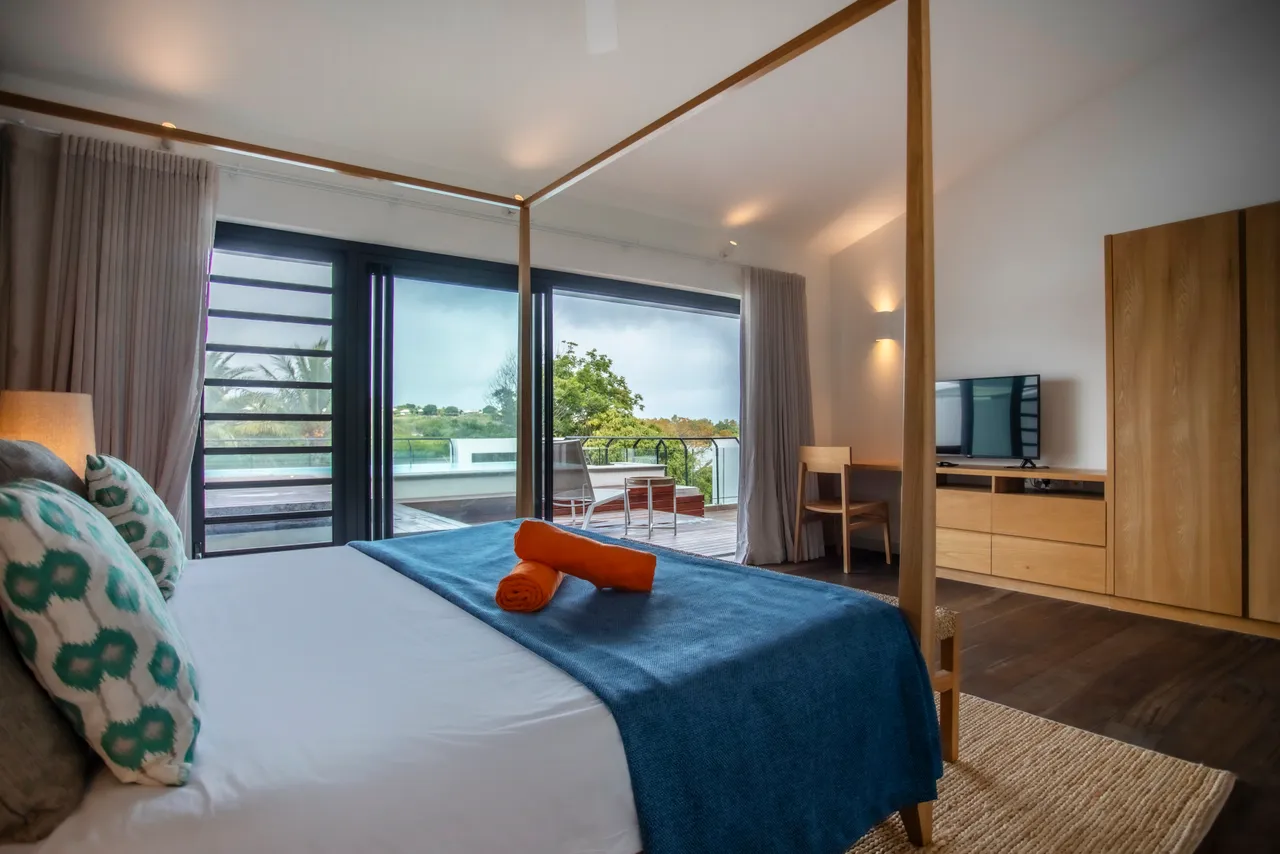 Chambre avec lit à baldaquin et accès à la terrasse en bois.  
Baies vitrées offrant une vue sur la piscine privée.  
Mobilier en bois clair ajoutant de la simplicité à l'espace.  
Tapis texturé apportant une touche de confort sous les pieds.  
Espace de rangement intégré pour une meilleure organisation.