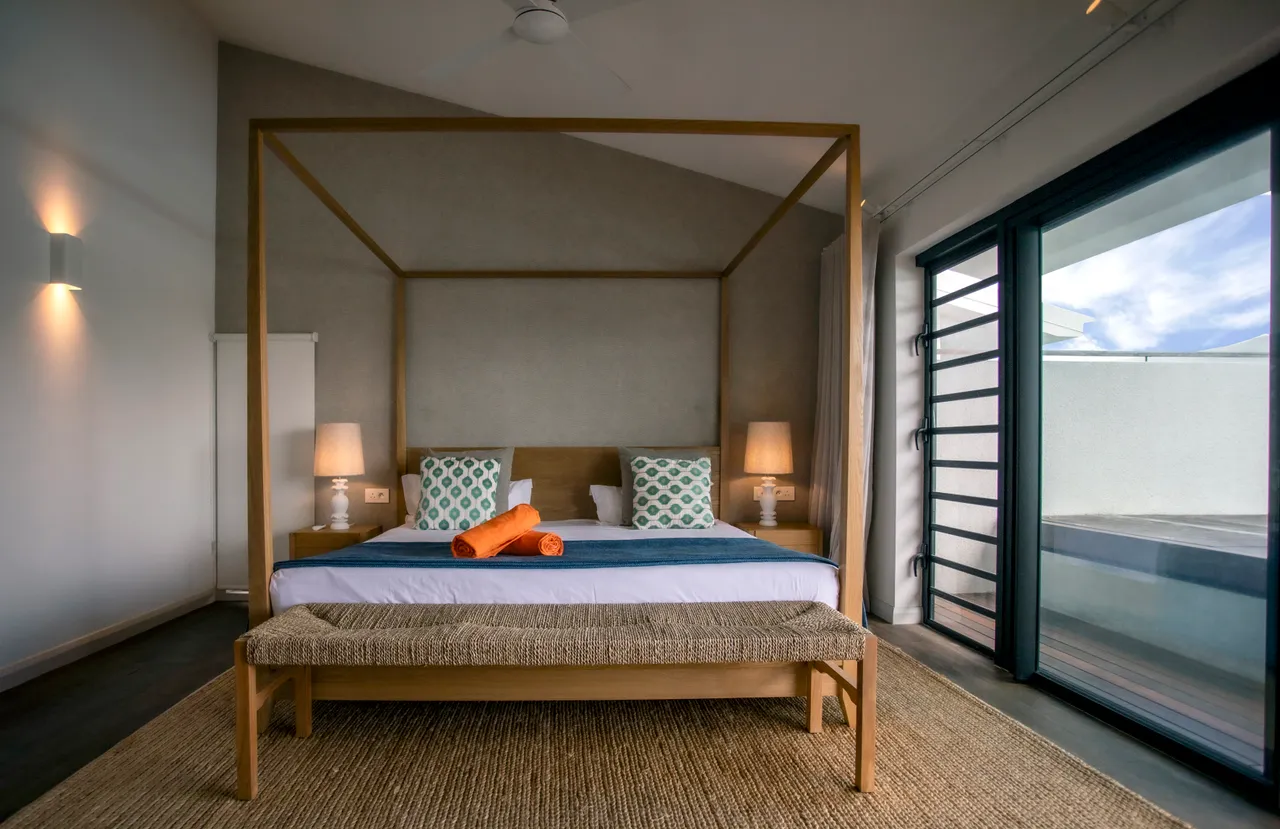 Chambre spacieuse avec un lit à baldaquin et accès à une terrasse.  
Luminaires encastrés apportant une luminosité adaptée à l'espace.  
Fenêtres panoramiques offrant une vue dégagée sur l'extérieur.  
Mobilier en bois clair créant une atmosphère tranquille.  
Tapis texturé ajoutant confort et chaleur au sol.