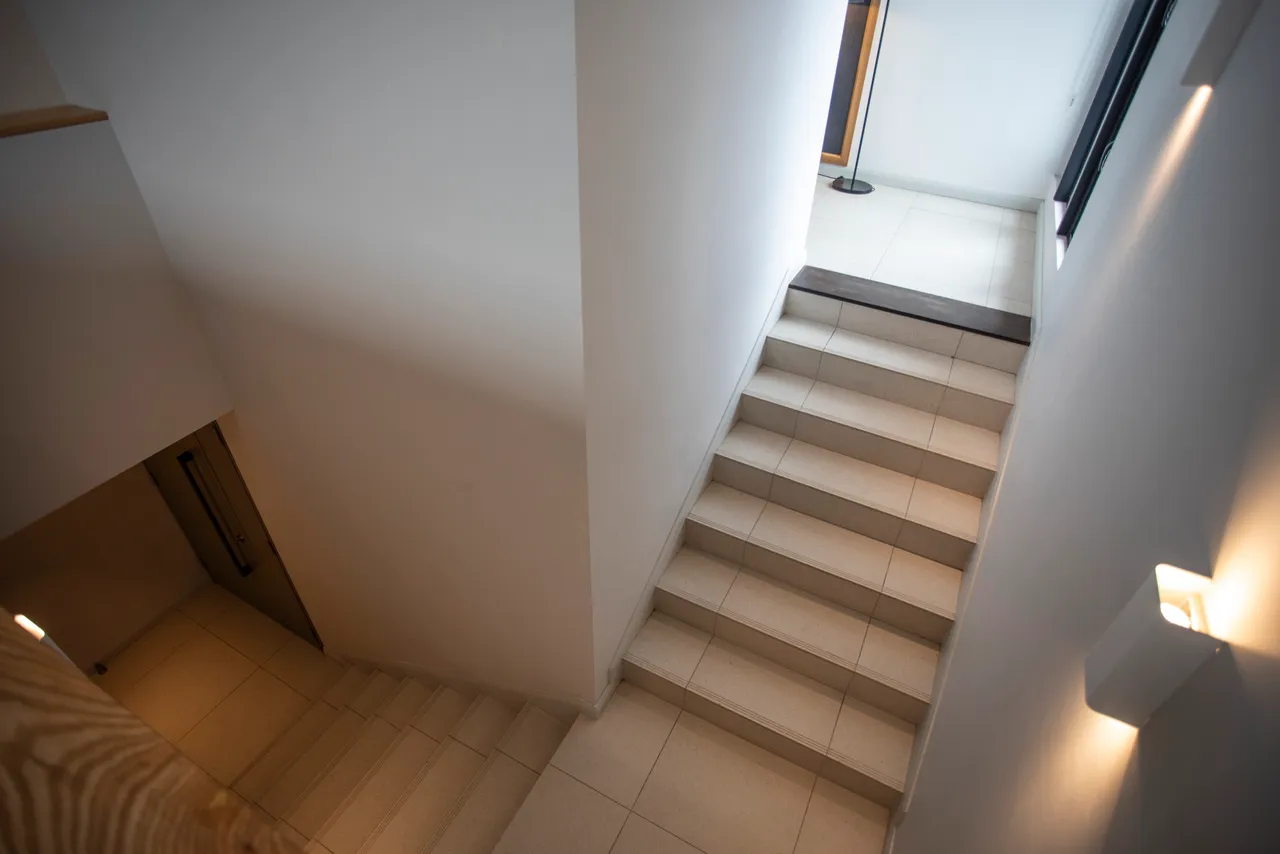 Escalier menant aux différents niveaux du penthouse.  
Murs blancs et lumière naturelle offrant une atmosphère ouverte.  
Marches carrelées facilitant la circulation entre les espaces.  
Éclairage intégré mettant en valeur la structure de l'escalier.  
Vue dégagée sur le reste du penthouse depuis le haut.