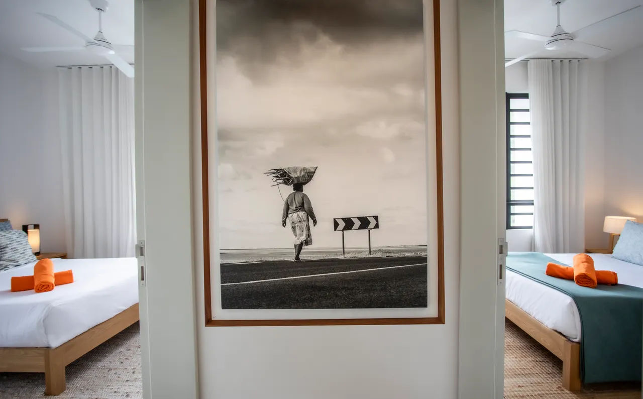 Photographie en noir et blanc encadrée créant un point focal dans l'espace.  
Sol en jute ajoutant une texture naturelle au couloir.  
Lits jumeaux avec linge de lit coordonné pour un style harmonieux.  
Éclairage doux sur les murs blancs pour une atmosphère apaisante.  
Rideaux occultants assurant intimité et obscurité dans