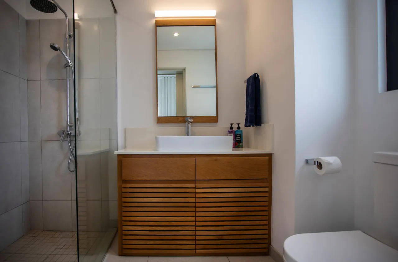 Salle de bains avec meuble en bois et plan vasque en surface claire.  
Douche moderne avec une paroi en verre et robinetterie contemporaine.  
Miroir sobre au-dessus du lavabo reflétant la lumière ambiante.  
Rangements intégrés offrant un espace pratique pour les produits.  
Carrelage neutre au sol apportant simplicité et facilité d'entretien.