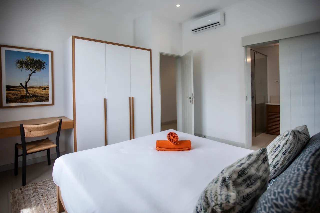 Chambre avec lit king-size et bureau intégré pour un espace fonctionnel.  
Mur décoré d'une photographie apportant une touche artistique.  
Rangements discrets intégrés pour un environnement épuré.  
Luminaire au-dessus du lit offrant un éclairage doux et chaleureux.  
Fenêtre large laissant entrer la lumière naturelle dans la pièce.