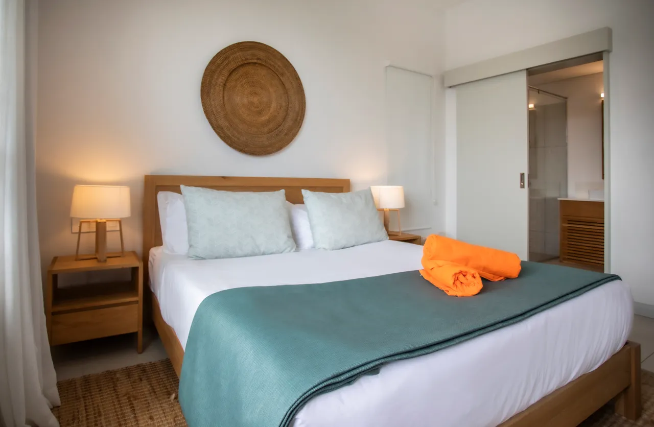 Chambre avec lit queen-size et linge de lit soigneusement disposé.  
Décoration murale en rotin ajoutant une touche naturelle à l'espace.  
Éclairage doux provenant des lampes de chevet de chaque côté du lit.  
Fenêtre laissant passer la lumière et offrant une vue sur l'extérieur.  
Espace de rangement pratique intégré au mobilier pour une meilleure organisation.