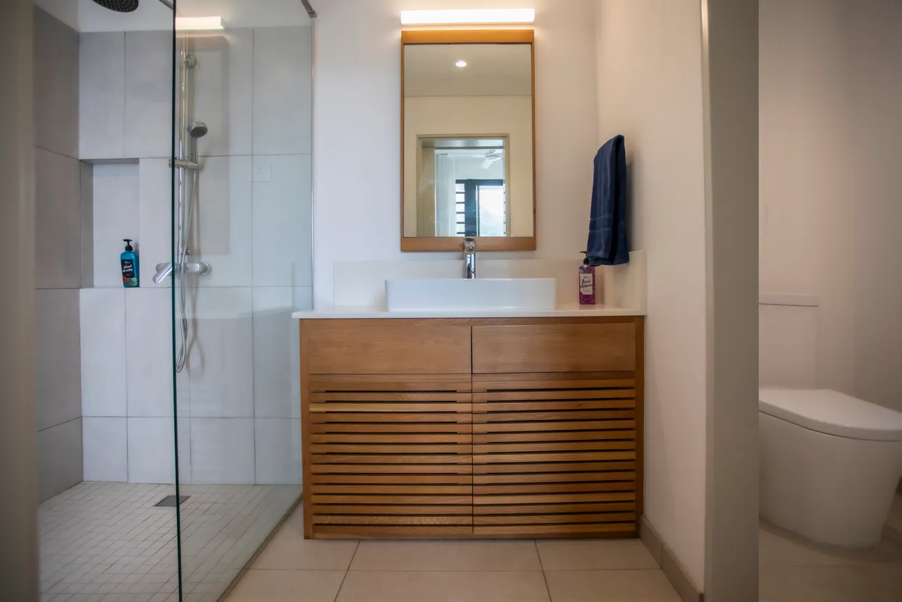 Tons neutres et meuble en bois dans la salle de bains.  
Douche moderne avec paroi en verre et robinetterie contemporaine.  
Miroir sobre réfléchissant la lumière pour un espace lumineux.  
Rangements intégrés offrant commodité pour les produits de toilette.  
Sol en carrelage facile d'entretien et au design épuré.