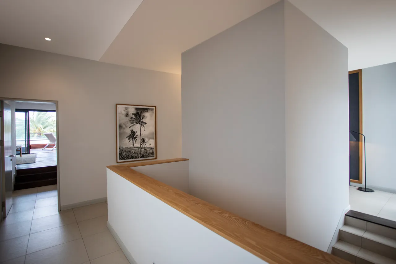 Galerie.  
Mur blanc avec une photographie en noir et blanc en exposition.  
Rampe en bois ajoutant une touche chaleureuse à l'espace.  
Accès direct à la terrasse avec vue sur la piscine.  
Éclairage encastré créant une ambiance lumineuse dans la galerie.