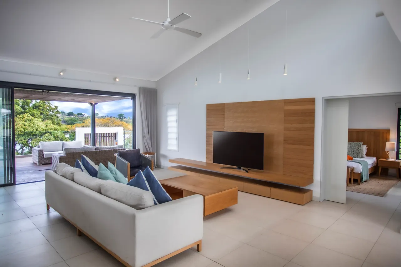 Espace de vie spacieux avec lumière naturelle et vue sur l'extérieur.  
Canapé moderne et table basse en bois créent un environnement fonctionnel.  
Télévision murale intégrée pour un divertissement accessible.  
Accès direct à la terrasse, prolongeant l'espace de vie.  
Ventilateur de plafond ajoutant confort et circulation d'air.