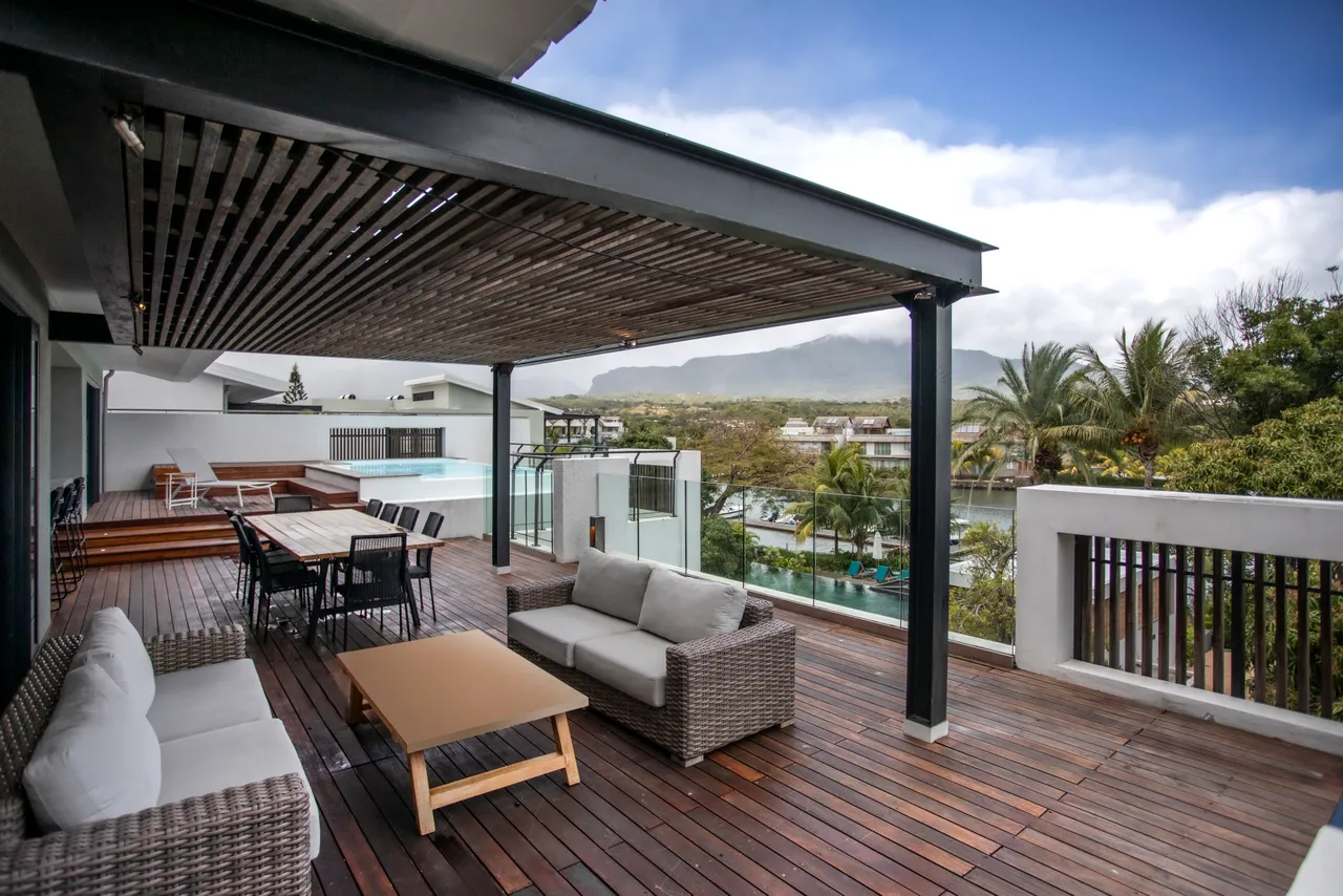 Terrasse en bois avec coin salon offrant une vue apaisante sur la nature.  
Ombrière moderne créant une zone d'ombre pour des moments de détente.  
Mobilier extérieur en rotin pour un confort durant les journées ensoleillées.  
Espace bien aménagé pour organiser des repas en plein air.  
Piscine visible depuis la terrasse, ajoutant un élément d'eau