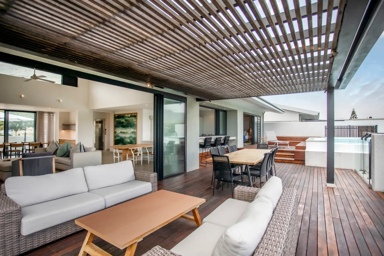 Terrasse spacieuse avec mobilier en rotin pour des moments de détente.  
Espace repas extérieur et piscine visible, favorisant les repas en plein air.  
Plafond en lattes de bois offrant une zone ombragée et esthétique.  
Végétation environnante ajoutant une touche naturelle autour de l'espace.  
Accès direct depuis le salon, facilitant la circulation entre