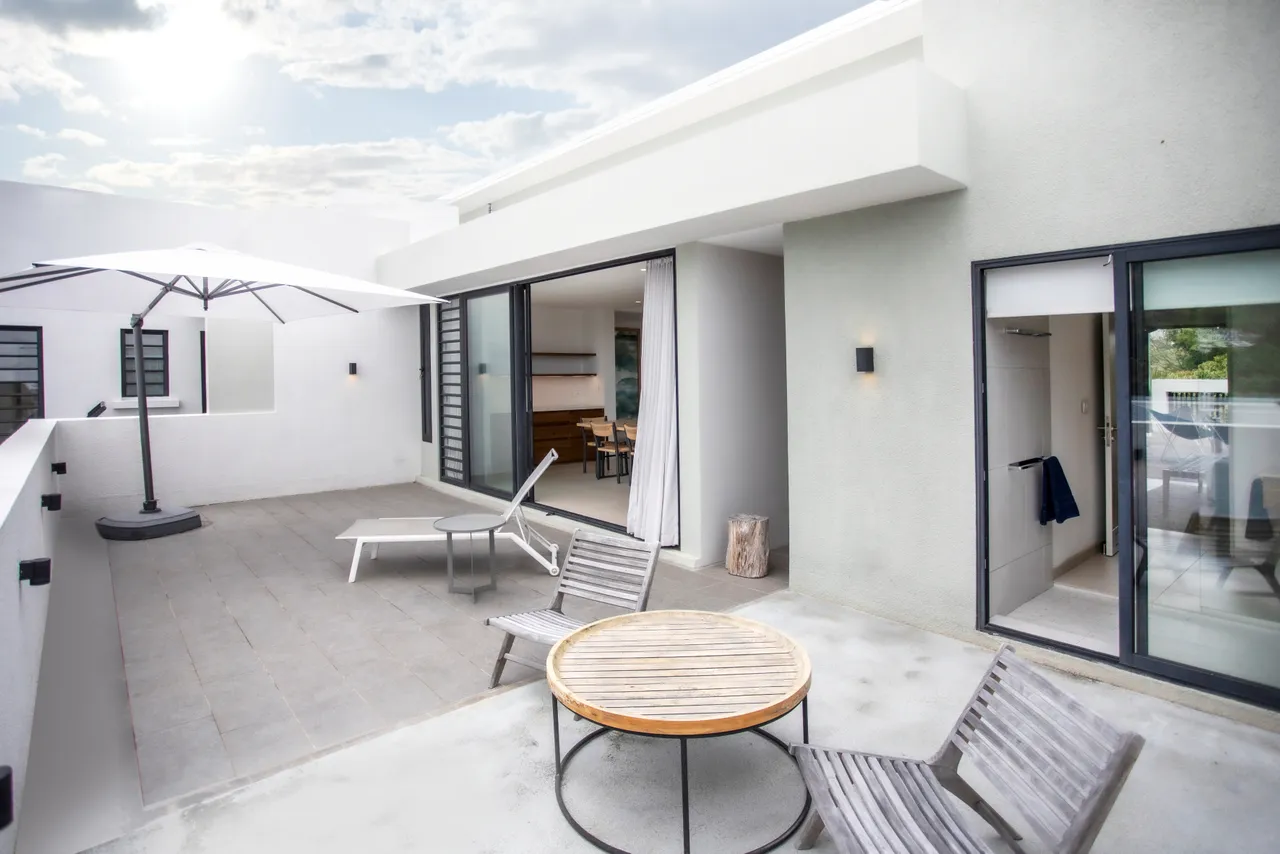 Terrasse spacieuse avec chaises longues pour se reposer au soleil.  
Mobilier en bois sur la terrasse pour un confort extérieur.  
Ombrelle blanche offrant de l'ombre lors des journées ensoleillées.  
Accès direct au salon par de grandes baies vitrées.  
Vue dégagée sur l'environnement naturel depuis l'espace extérieur.
