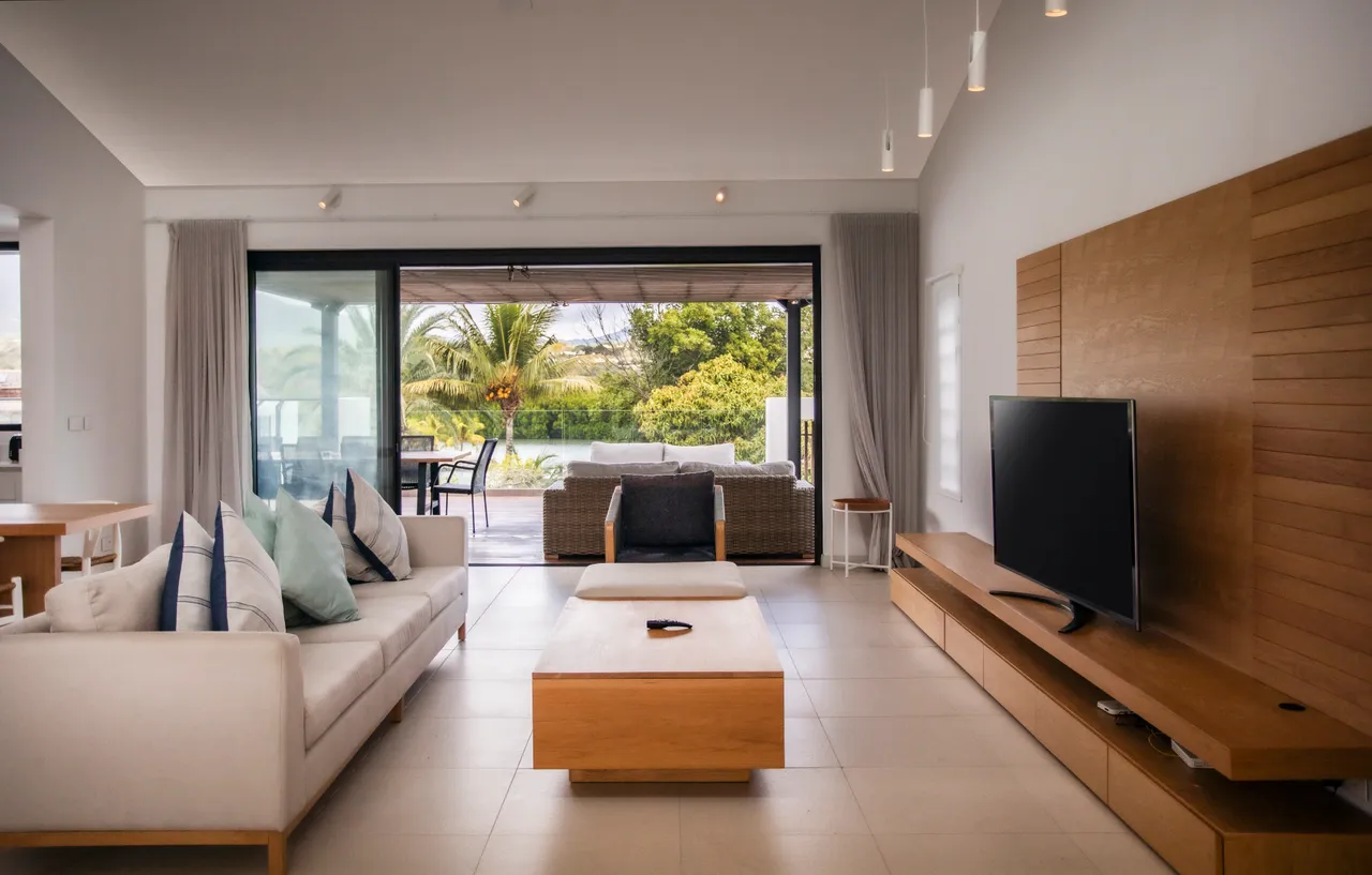 Tons clairs et mobilier en bois créent une ambiance chaleureuse.  
Salon lumineux avec accès direct à la terrasse extérieure.  
Écran TV mural intégré pour un divertissement à portée de main.  
Grandes baies vitrées offrant une vue sur le paysage environnant.  
Espace de vie fonctionnel avec canapé et table basse moderne.