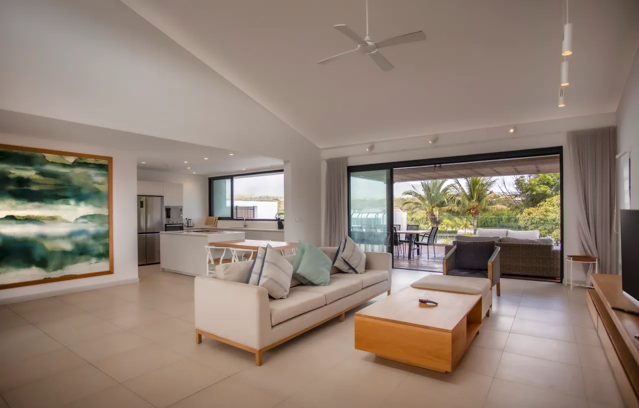 Sol en carrelage clair avec mobilier moderne et épuré.  
Grand canapé angulaire offrant confort et espace de détente.  
Table basse en bois ajoutant une touche naturelle à l'espace.  
Larges baies vitrées permettant une vue sur la terrasse extérieure.  
Éclairage intégré créant une ambiance agréable dans le salon.