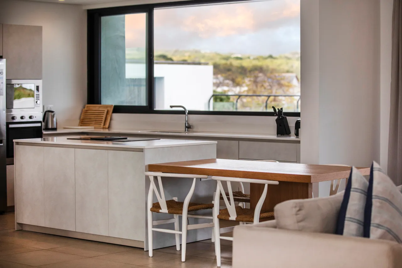 Cuisine moderne avec un îlot central et des appareils intégrés.  
Espace repas lumineux avec des chaises blanches et une table en bois.  
Larges fenêtres laissant entrer la lumière naturelle dans la pièce.  
Agencement fonctionnel facilitant la préparation des repas.  
Mobilier épuré et contemporain pour un environnement pratique.