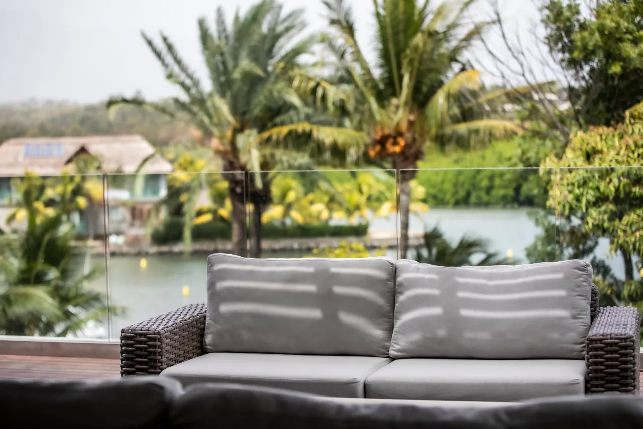 Espace extérieur avec canapé en rotin, offrant une vue sur la végétation.  
Végétation environnante créant un cadre naturel apaisant autour du salon.  
Terrasse spacieuse avec une ambiance détendue, idéale pour se reposer.  
Accès direct à la piscine depuis l'espace extérieur, facilitant les baignades.  
Luminosité naturelle accentuée par la