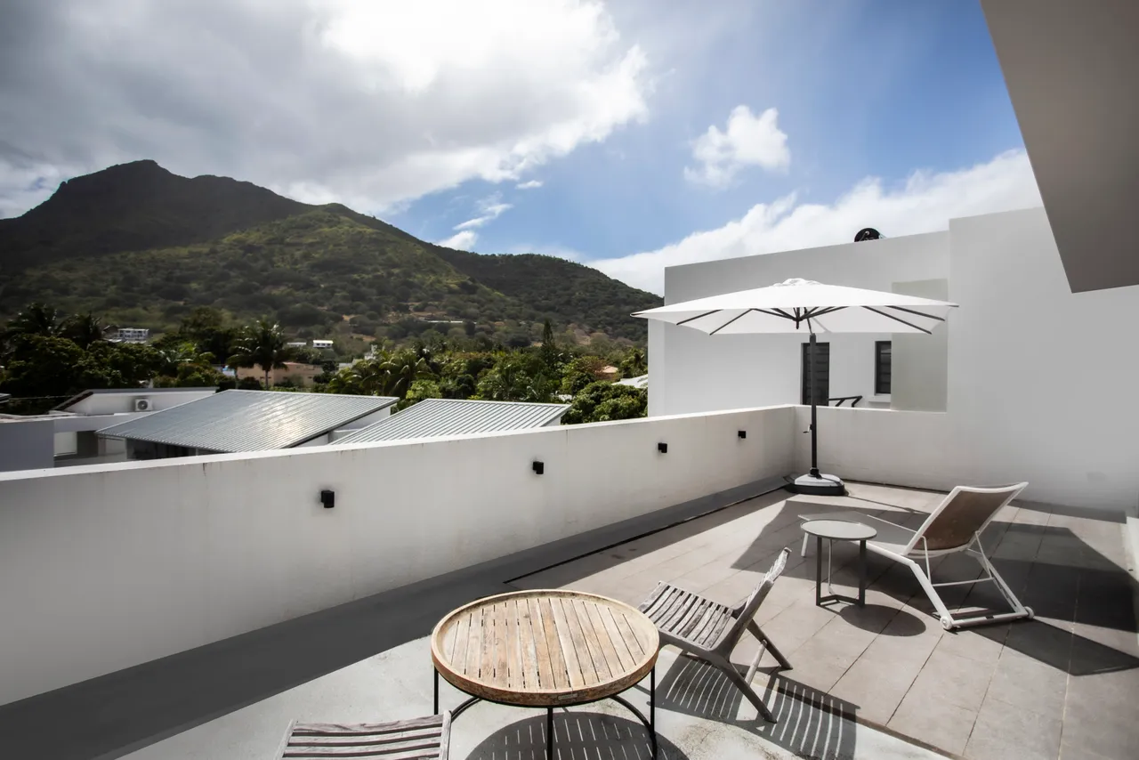 Terrasse en carrelage avec table ronde et parasol pour des repas extérieurs.  
Chaises longues en métal offrant un espace pour se relaxer au soleil.  
Vue dégagée sur la montagne, ajoutant une dimension naturelle à l'espace.  
Espace extérieur spacieux permettant de profiter du climat de l'île.  
Ambiance sereine grâce à la disposition des meubles et à la