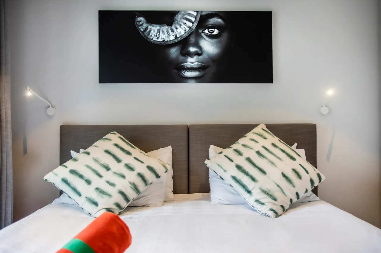 Textures douces et motifs géométriques sur les coussins du lit.  
Décor mural artistique ajoutant une touche de caractère à la chambre.  
Éclairage minimaliste avec lampes murales intégrées au design.  
Lit king-size avec linge de lit blanc favorisant la simplicité.  
Rangement discret avec des espaces optimisés sous la tête de lit.  
Ambiance reposante grâce