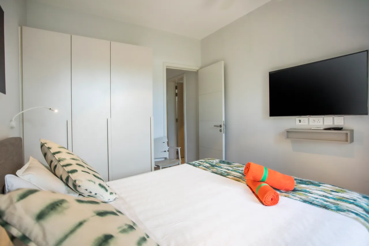 Chambre équipée d'une télévision murale pour le divertissement.  
Rangement spacieux avec des armoires blanches discrètes.  
Lit double avec coussins aux motifs variés et colorés.  
Espace de travail fonctionnel intégré à l'aménagement.  
Douche moderne dotée d'un carrelage soigné et élégant.  
Ambiance lumineuse grâce à des fenêtres offrant