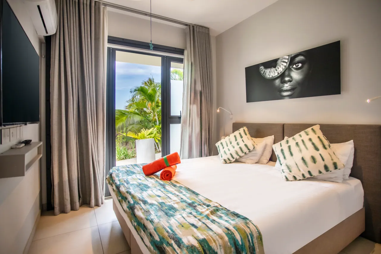 Tons neutres et textures douces dans la chambre à coucher.  
Grandes fenêtres offrant une vue sur la végétation environnante.  
Éclairage discret avec lampes murales intégrées au design.  
Coussins colorés ajoutent du confort au lit double.  
Télévision murale pour un divertissement accessible.  
Rangement optimisé avec armoires blanches intégrées.
