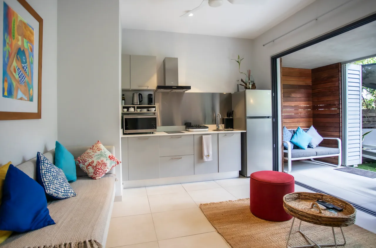 Cuisine moderne avec des appareils intégrés et un comptoir fonctionnel.  
Coin salon lumineux avec un accès direct à la terrasse.  
Mobilier contemporain disposé pour optimiser l'espace de vie.  
Coussins colorés ajoutent une touche de confort au banc.  
Vue sur l'extérieur depuis les larges ouvertures du studio.  
Espace bien agencé pour favoriser la convivialité.