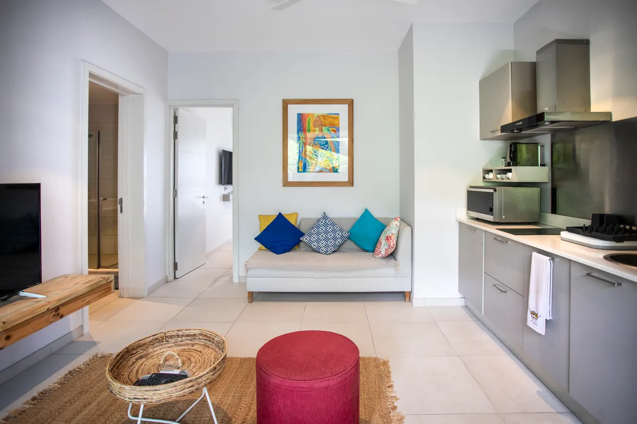 Coin salon avec banc confortablement garni de coussins colorés.  
Cuisine moderne équipée d'un plan de travail fonctionnel.  
Table basse en osier ajoutant une touche naturelle à l'espace.  
Éclairage naturel grâce à des fenêtres offrant une vue sur l'extérieur.  
Décoration murale colorée ajoutant du caractère à l'intérieur.  
Rangement intégré permettant d'optimiser