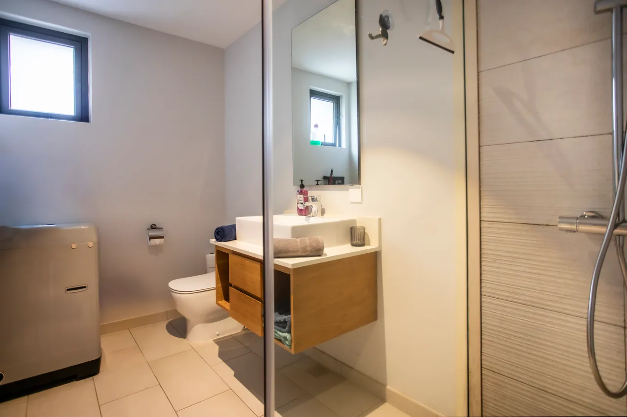 Salle de bain avec un meuble sous vasque en bois et miroir intégré.  
Douche spacieuse avec panneaux de verre et carrelage sobre.  
Éclairage fonctionnel au-dessus du miroir pour un usage pratique.  
Linge de bain soigneusement disposé sur un espace de rangement.  
Équipement moderne pour le confort au quotidien dans la salle de bain.  
Coin buanderie